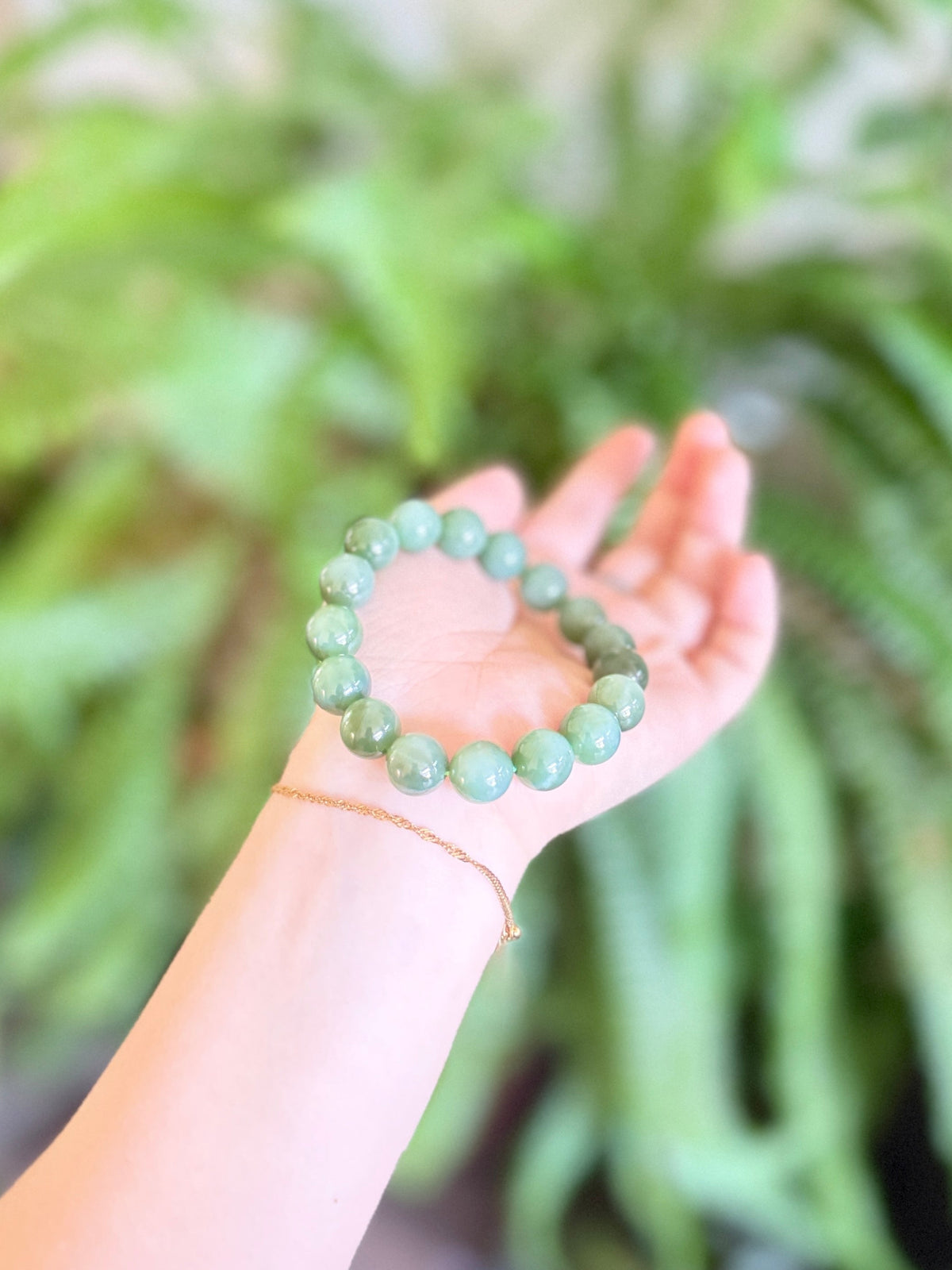 A person's hand holding a light green beaded bracelet, with a thin gold chain bracelet also visible on the wrist, set against a soft-focus background of green plants.