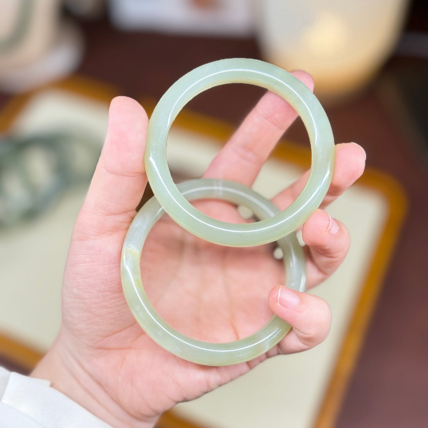 A hand holding two light green jadeite bangles, with a softly blurred indoor background.