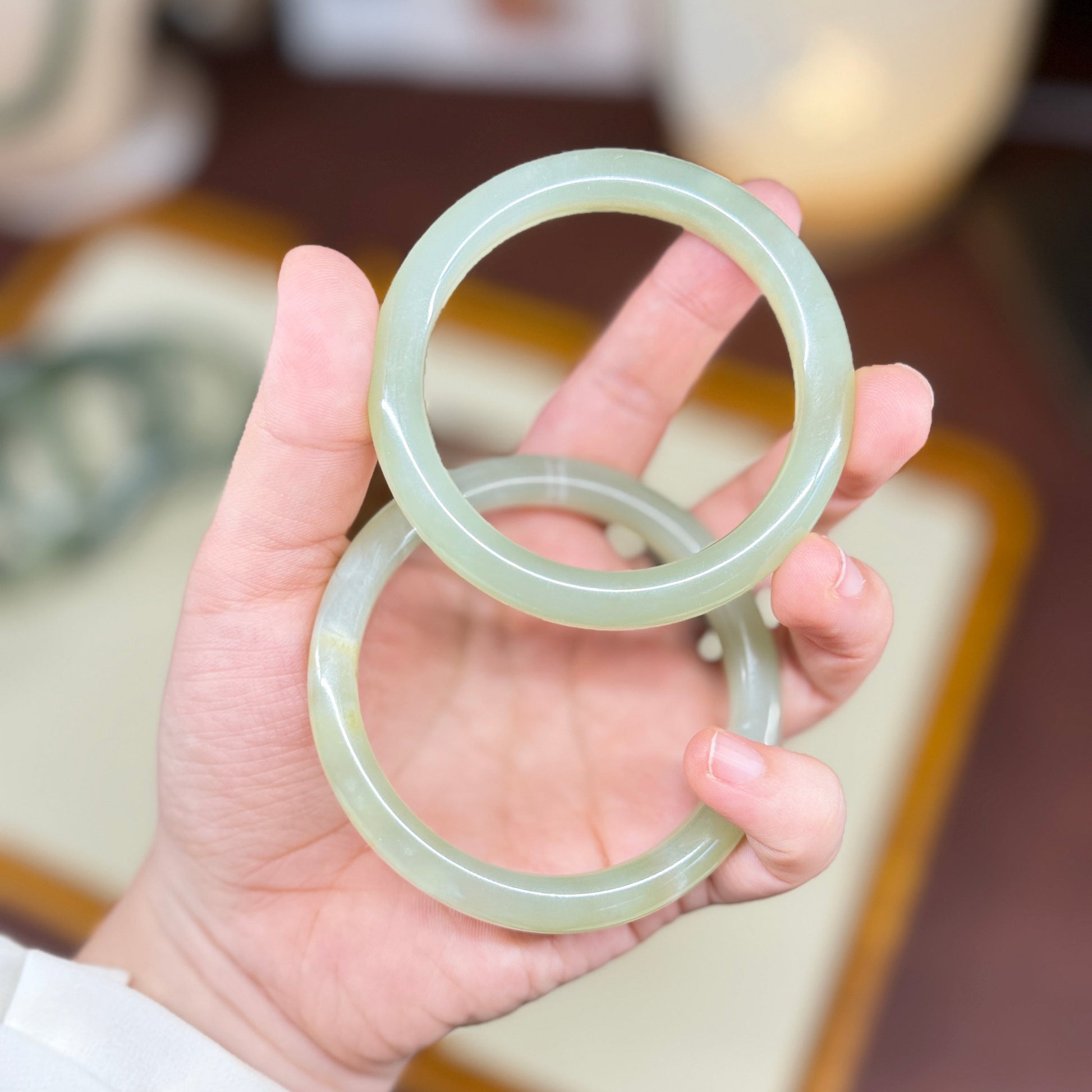 A hand holding two light green jadeite bangles, with a softly blurred indoor background.
