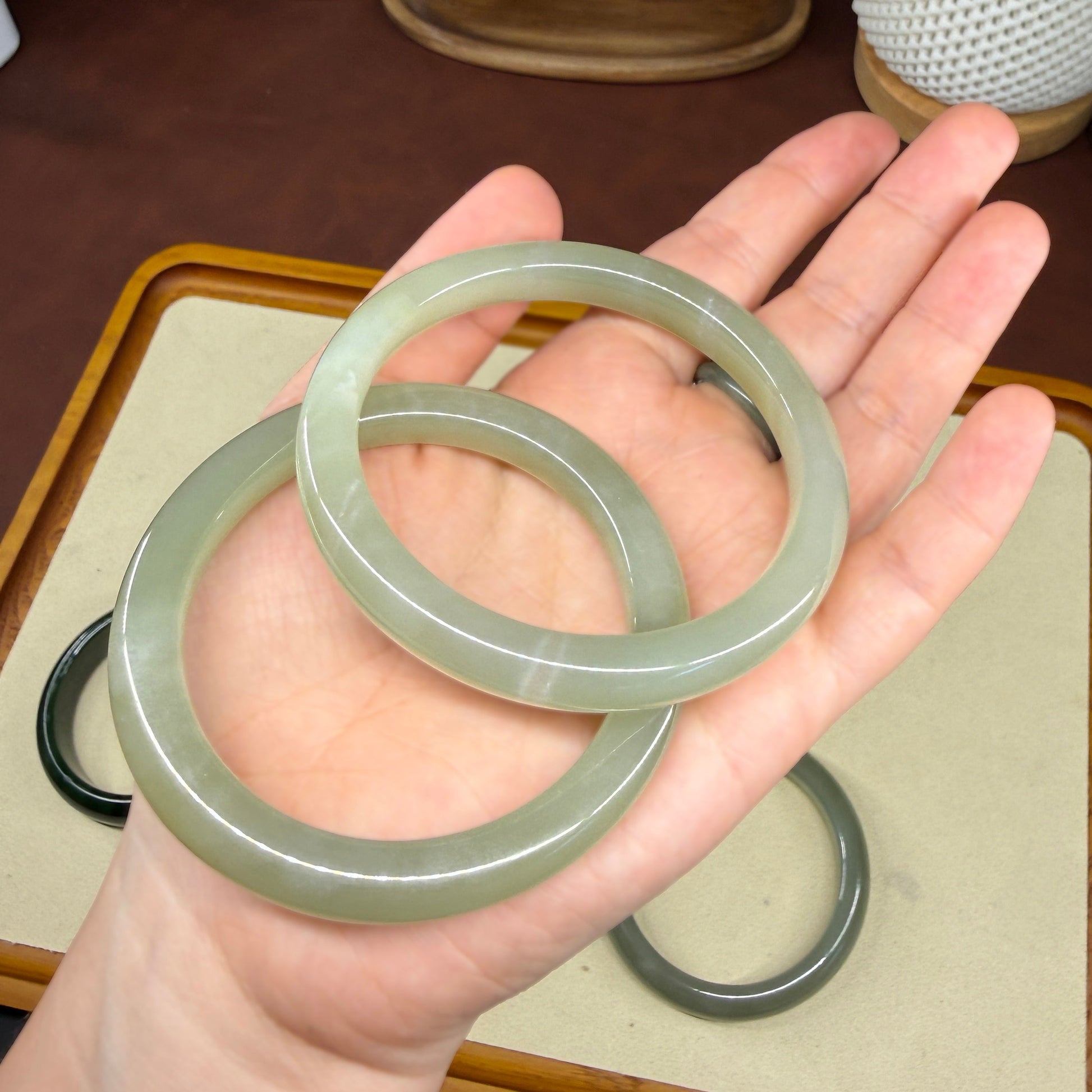 A person's hand holding two light green jadeite bangles, with more bangles displayed on a tray in the background of a styled indoor setting.