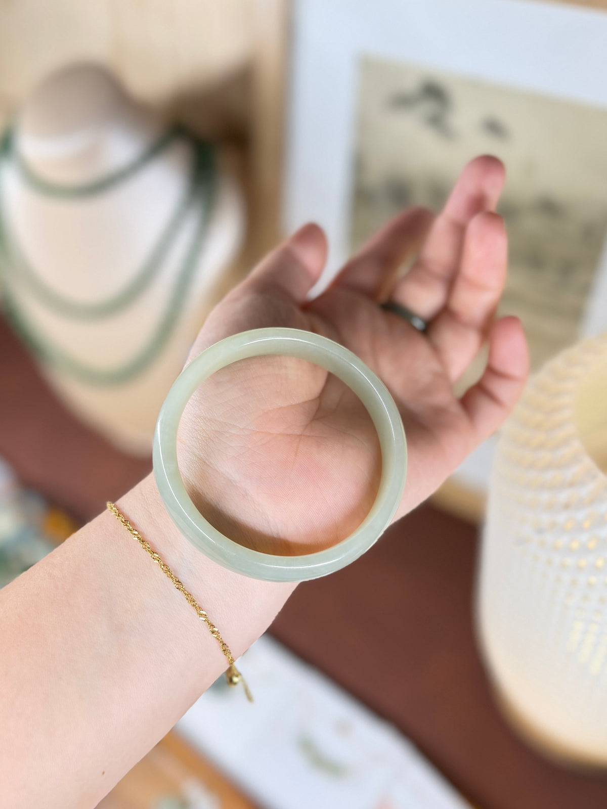 A translucent pale green jadeite bangle bracelet being worn on a person's wrist, with a delicate gold chain bracelet also visible. The hand is held up against a softly blurred indoor background with warm lighting.