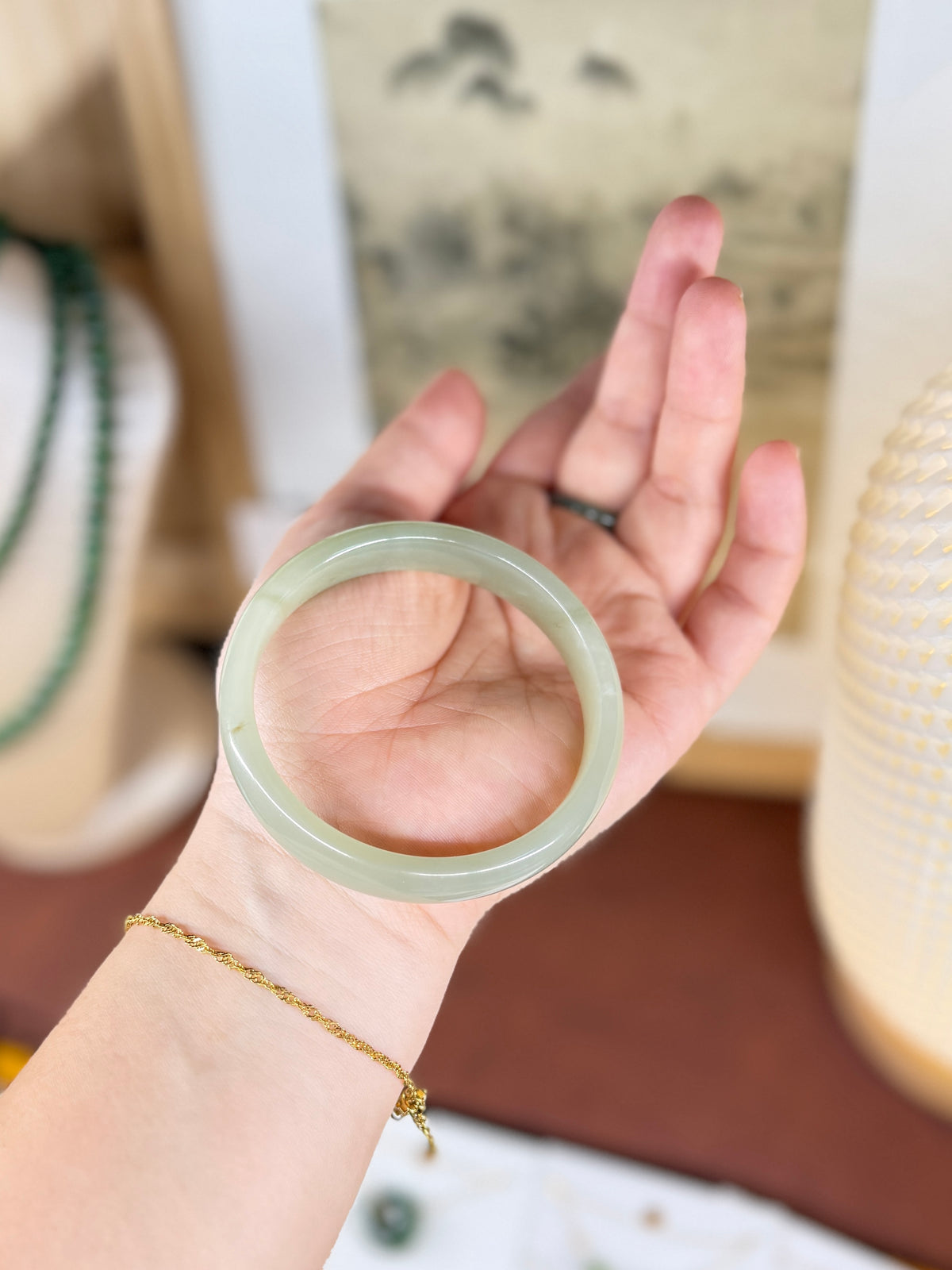 A translucent pale green jade bangle bracelet held in a person's hand, with a delicate gold chain bracelet on the wrist and a blurred indoor background.