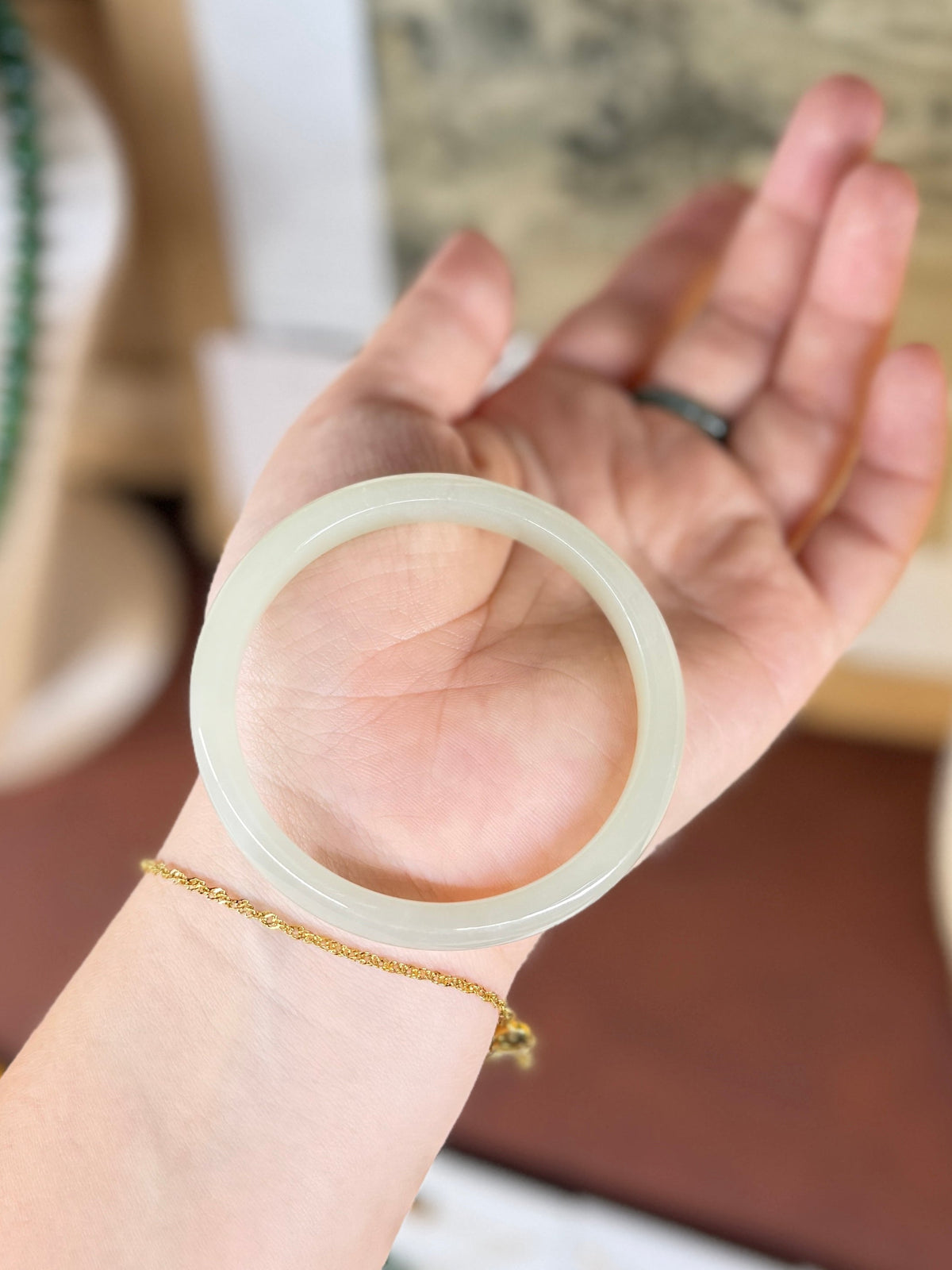 A translucent pale green jade bangle held in a person's hand, with a delicate gold chain bracelet on the wrist and a dark ring on the finger, set against a softly blurred indoor background.