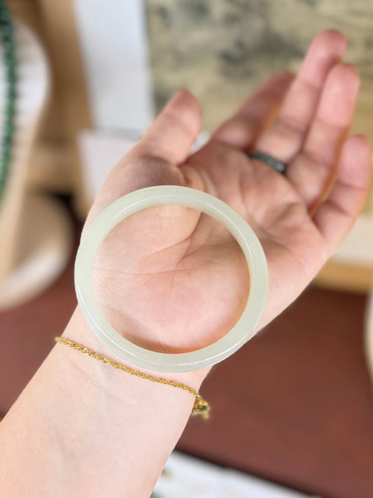 A translucent pale green jade bangle held in a person's hand, with a delicate gold chain bracelet on the wrist and a dark ring on the finger, set against a softly blurred indoor background.