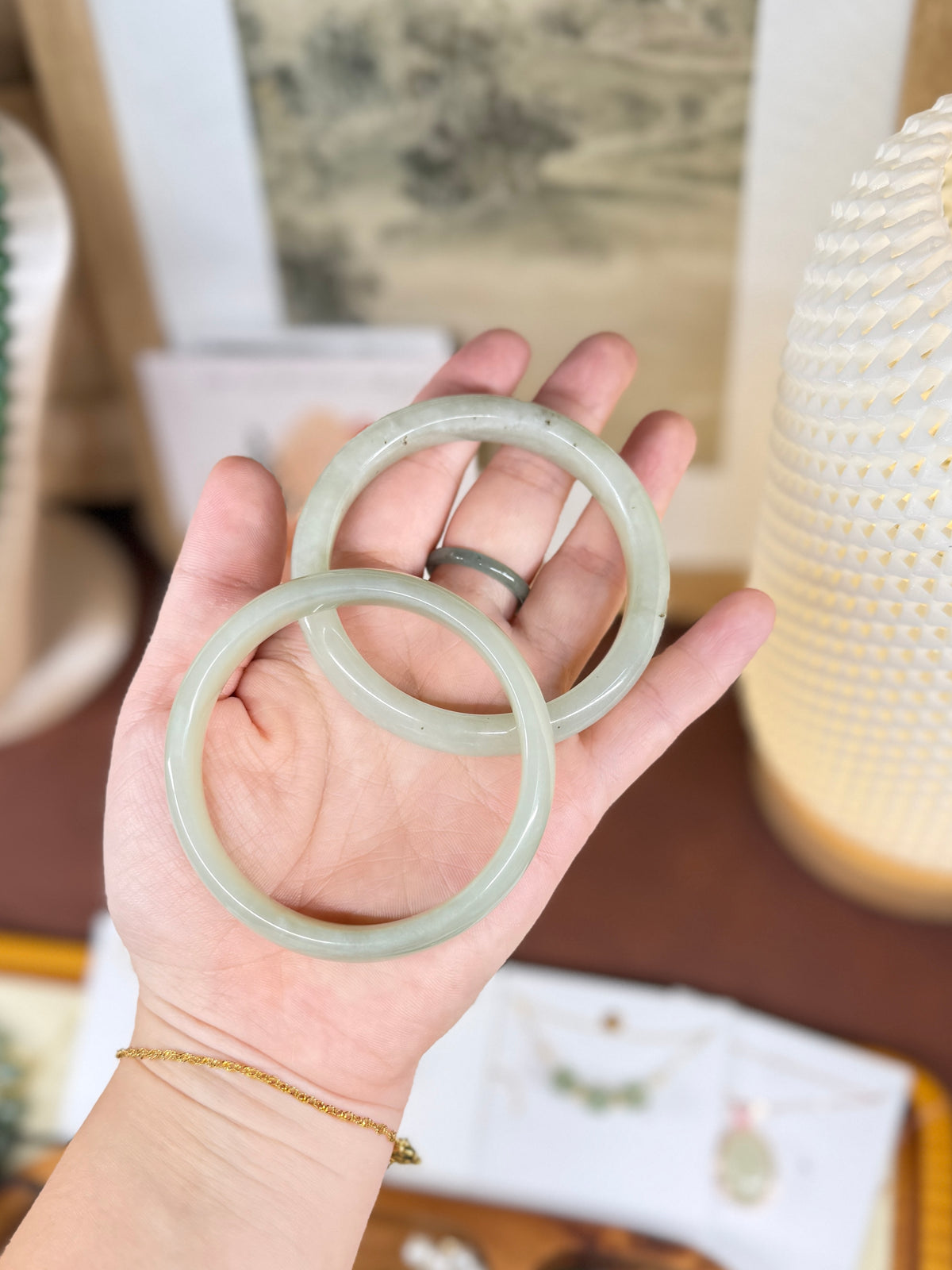 A hand holding two light green jadeite bangles, with a gold ring on the index finger, set against a softly blurred background of home decor.