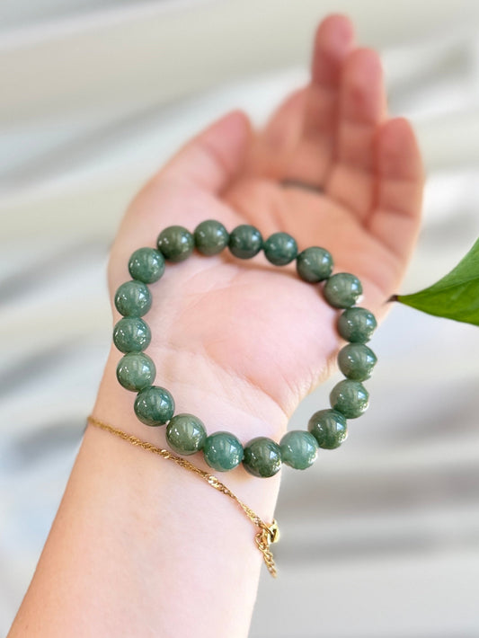 A green beaded bracelet and a delicate gold chain bracelet with a small charm, displayed on a person's wrist and palm against a soft, blurred white background with a green leaf visible on the right.
