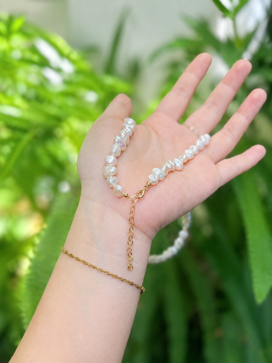 A white baroque pearl necklace with a gold clasp and extender chain, draped over an open hand. A matching gold chain bracelet is worn on the wrist, set against a soft-focus background of green foliage.