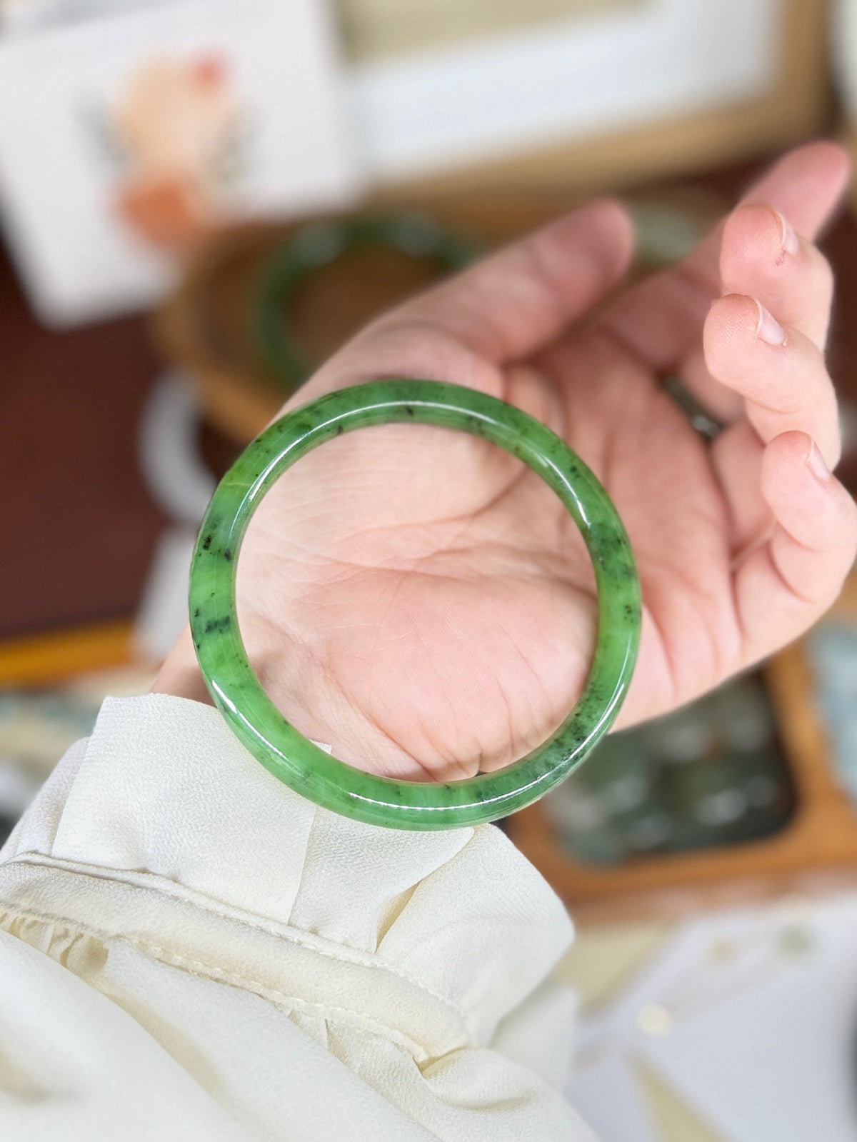 A hand holding a green jadeite bangle bracelet, with other jewelry items and a warm-toned background visible.