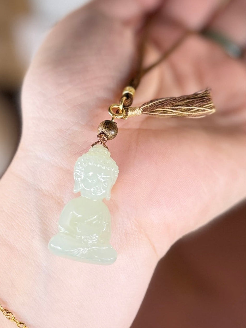 A close-up of a hand holding a small carved pendant necklace. The pendant is made of pale translucent jade-like material and hangs from a gold-toned cord with a tassel and small gold beads.