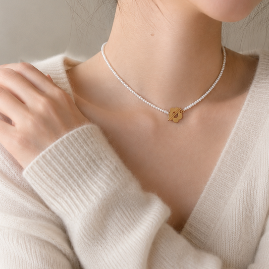 A close-up of a model wearing a delicate white pearl choker necklace with a small gold flower-shaped pendant, paired with a cream-colored ribbed knit sweater.